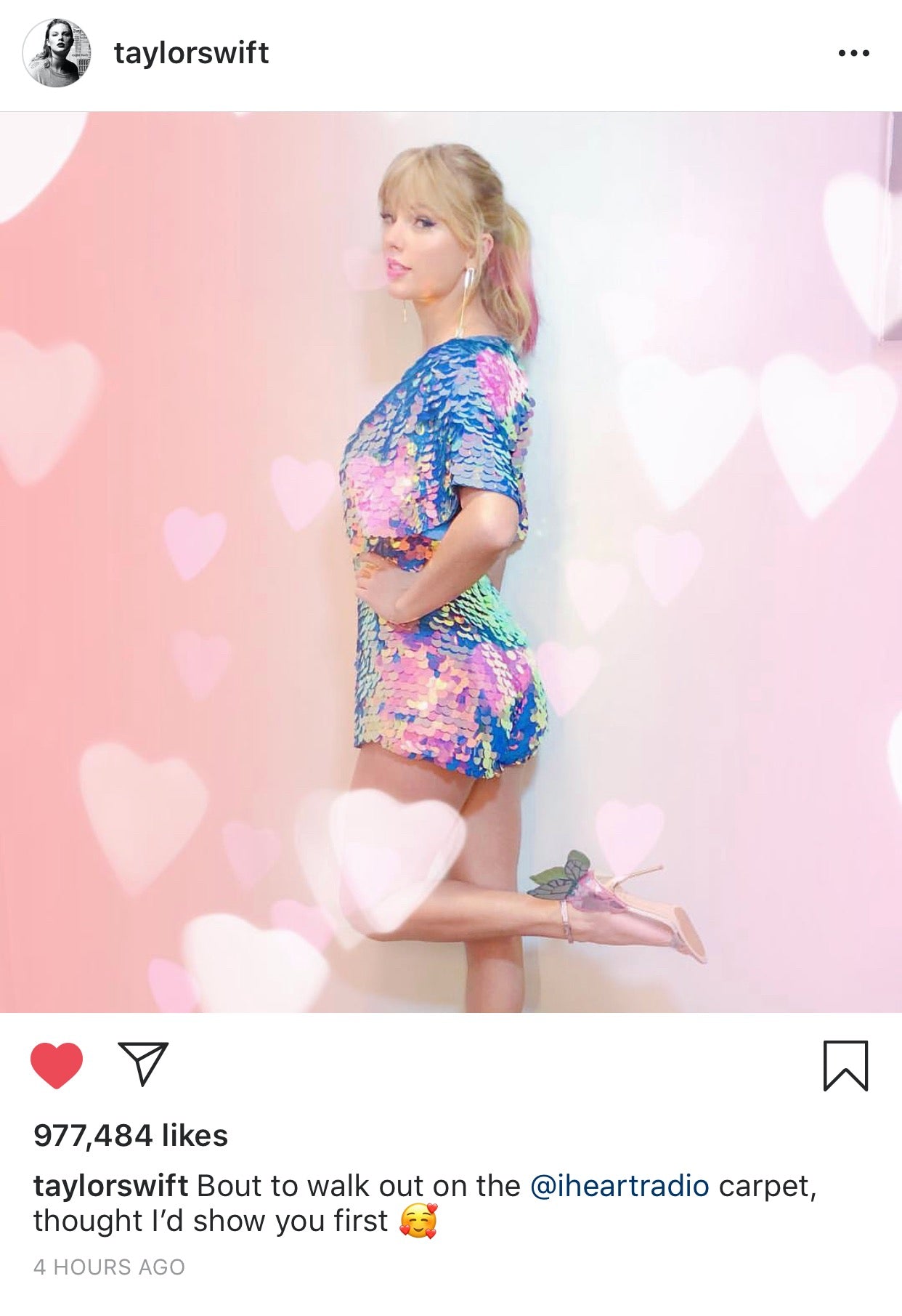 An Instagram post from Taylor Swift wearing the Rosa Bloom Orchid Mella playsuit to the I heart radio awards ceremony. She is standing on one leg shoeing off her butterfly heeled shoes and iridescent blue, pink and purple sequin outfit with commanding cape sleeves and shorts.