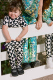 A cropped image of a small child with short dark hair who is standing on a white fence. Cropped from the image is older children wearing kids sequin festival and party leggings and black and white tri-print leggings. The little girl is wearing black and white geometric and triangle pair of childrens leggings in the Rosa Bloom tri-print design with a matching childrens t-shirt. . 