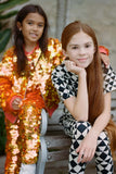 Two children with long hair looking at the camera in an outdoor park setting.  One is wearing a fox orange and yellow shimmering sequin festival kids t-shirt and leggings, with matching childrens sequin bomber jacket. The other is wearing a black and white geometric and triangle pair of Childrens leggings with a matching short sleeve black and white geometric tri-print t-shirt. 