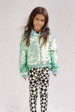 A child with long dark hair looking at the camera against a white wall.  She is wearing a mint green, chameleon bomber jacket. A mix of cream and green shimmering sequin festival jacket, with co-ordinating black and white geometric and triangle pair of childrens leggings in the Rosa Bloom tri-print design. 