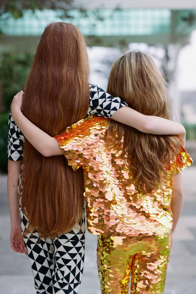 Two children with long hair with their backs to us in an outdoor park setting. The two children have their arms around each other. One is wearing a fox orange and yellow shimmering sequin festival kids t-shirt and leggings. The other is wearing a black and white geometric and triangle pair of Childrens leggings with a matching short sleeve black and white geometric tri-print t-shirt.