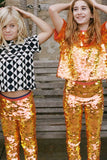 Two children who look like they are having fun in an outdoor park setting, standing in front of a park bench. They are wearing a mix of orange, gold and yellow large round sequin festival kids childrenswear in leggings and t-shirt, and a geometric graphic black and white Tri-print t-shirt by Rosa Bloom.