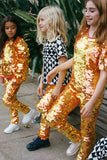 Three children in a brightly lit outdoor walled garden space with tropical plants. They are all wearing Rosa Bloom fox m orange, gold and yellow, shimmering sequin clothing. They are wearing the fox sequin leggings, sequin childrens t-shirts and matching festival sequin bomber jacket. The middle child is dressed in Childrenswear Rosa Bloom tri-print in black and white.