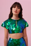 A woman with brown hair, wearing a festival sequin cape made with small and large round holographic Rosa Bloom sequins. The Emerald sequin cape, sitting neatly over her shoulders in this Inti design glistens in the light, creating a mix of shimmering colours of green and blue. The model is also wearing a matching sequin vest top and sparkly leggings.