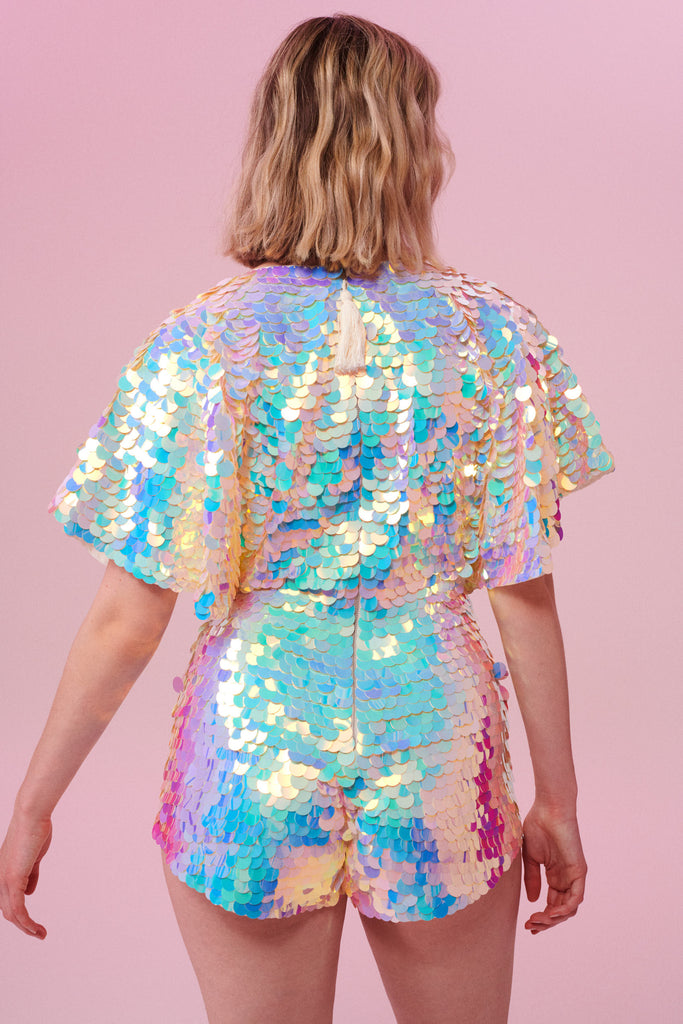 A model with short blonde hair with her back to the camera wearing a Rosa Bloom sequin Mella romper playsuit in the Opal design. A mix of white, pinks and blue sequins shimmer in the light showing the shorts and commanding cape sleeved festival outfit.  The all over iridescent disc sequins, as worn by Taylor Swift shimmers in the light.