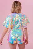 A model with short blonde hair with her back to the camera wearing a Rosa Bloom sequin Mella romper playsuit in the Opal design. A mix of white, pinks and blue sequins shimmer in the light showing the shorts and commanding cape sleeved festival outfit.  The all over iridescent disc sequins, as worn by Taylor Swift shimmers in the light.  