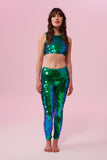 A woman with long brown hair wearing iridescent blue and green Indus leggings covered in large round holographic Rosa Bloom sequins. The sequins glisten, creating a mix of shimmering colours make this sequin glow. The model is also wearing a matching stretchy sequin vest top, in matching colours. This Emerald sequin outfit looks like it is glowing. 