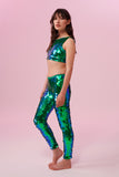 A woman with long brown hair wearing iridescent blue and green Indus leggings covered in large round holographic Rosa Bloom sequins. The sequins glisten, creating a mix of shimmering colours make this sequin glow. The model is also wearing a matching stretchy sequin vest top, in matching colours. This Emerald sequin outfit looks like it is glowing. 