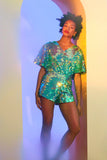 A model with short brown hair wearing a Rosa Bloom sequin Mella romper playsuit in the chameleon design. A mix of greens, purples and blue sequins shimmer in the light showing the shorts and commanding cape sleeved outfit.  The iridescent disc sequins, as worn by Taylor Swift shimmers in the light.  