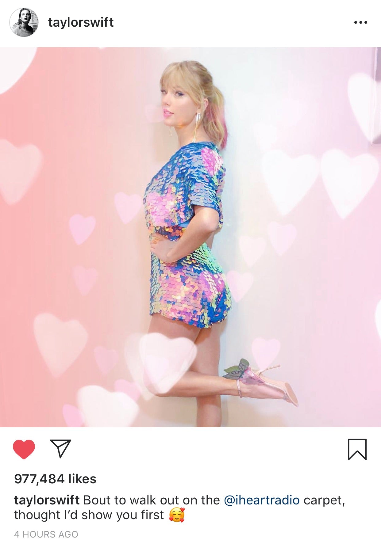An Instagram post from Taylor Swift wearing the Rosa Bloom Orchid Mella playsuit to the I heart radio awards ceremony. She is standing on one leg shoeing off her butterfly heeled shoes and iridescent blue, pink and purple sequin outfit with commanding cape sleeves and shorts. 
