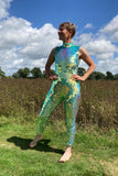 A woman with short hair, wearing an all-in-one sequin stretchy festival jumpsuit made with large round holographic Rosa Bloom sequins. The woman standing outside on a sunny day in a field is wearing the Iris jumpsuit. The sequins glisten all over in this chameleon colour way, creating a mix of shimmering colours of soft mint green and sage that sparkles in the light.