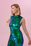 The top half of a woman with short blonde hair, wearing an all-in-one sequin stretchy festival jumpsuit made with large round holographic Rosa Bloom sequins. The woman  is wearing the Emerald jumpsuit in front of a pink background. The sequins glisten all over in this emerald colour way, creating a mix of shimmering sequin colours of green and blue that sparkles in the light.