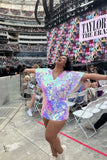 A photo of a woman at large stadium wearing a Rosa Bloom Opal Mella playsuit. She is standing with her arms out to the side with her head tilted back towards the sky looking very happy. She is wearing an iridescent blue, pink and purple sequin Rosa Bloom playsuit with commanding cape sleeves and shorts.