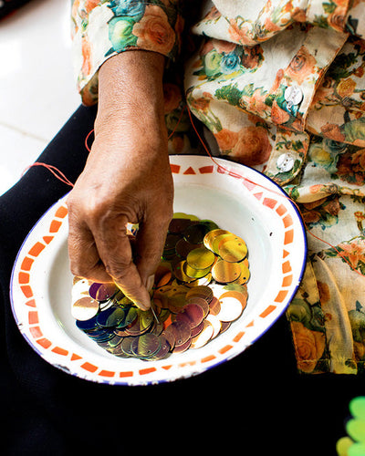 Person wearing a floral dress and holding a needle and thread with a bowl of colourful sequins on her lap