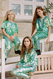 Three children with long hair in a brightly lit outdoor walled garden. The children are posing for the photo sitting on a white fence and park bench. They are all wearing Rosa Bloom chameleon mint green, shimmering sequin childrens t-shirt and matching festival sequin childrens Luna design leggings. 