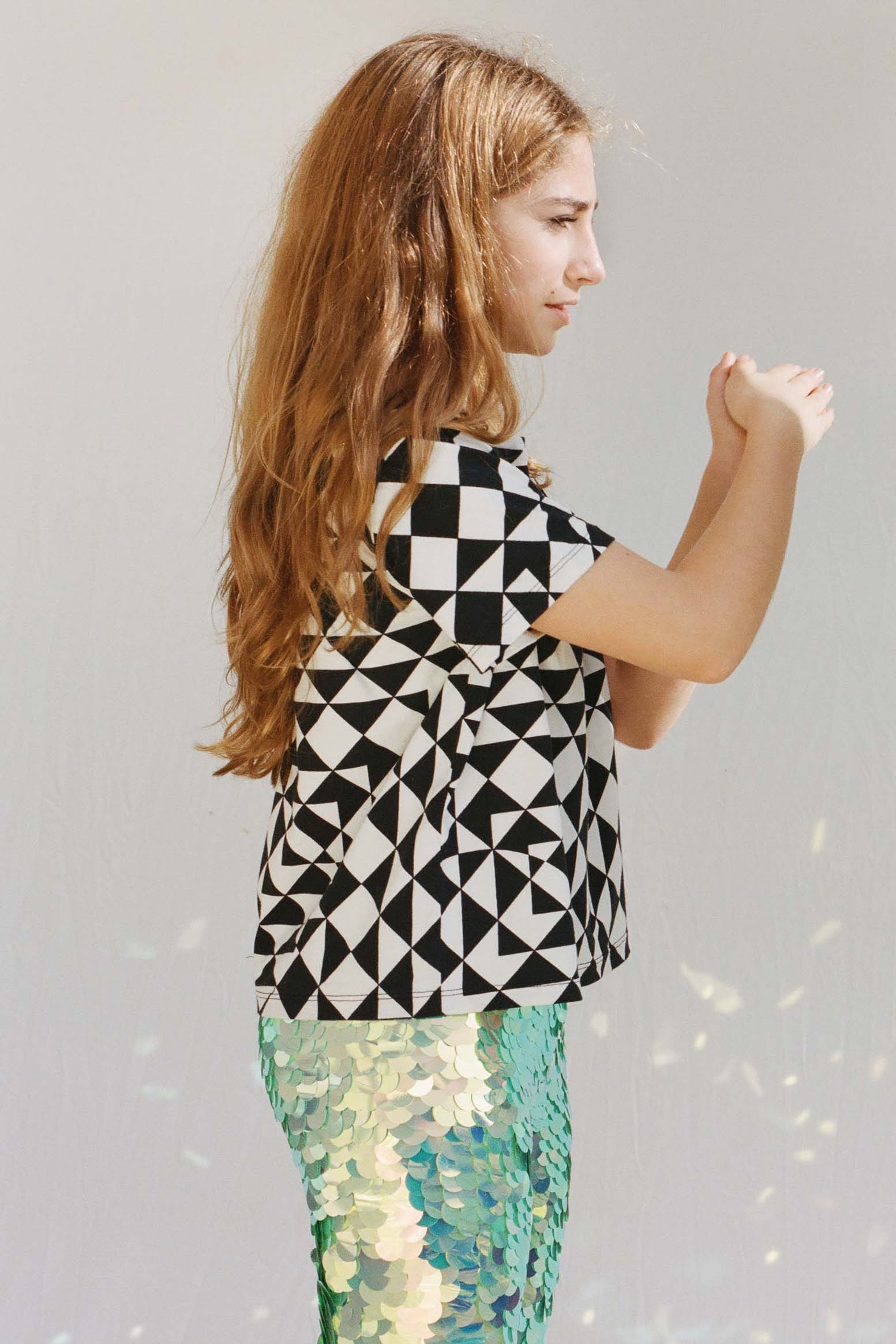 A girl with long hair standing side on to the camera in a brightly lit outdoor space wearing Rosa Bloom chameleon mint green children’s leggings, with contrasting bold graphic black and white tri-print t-shirt.