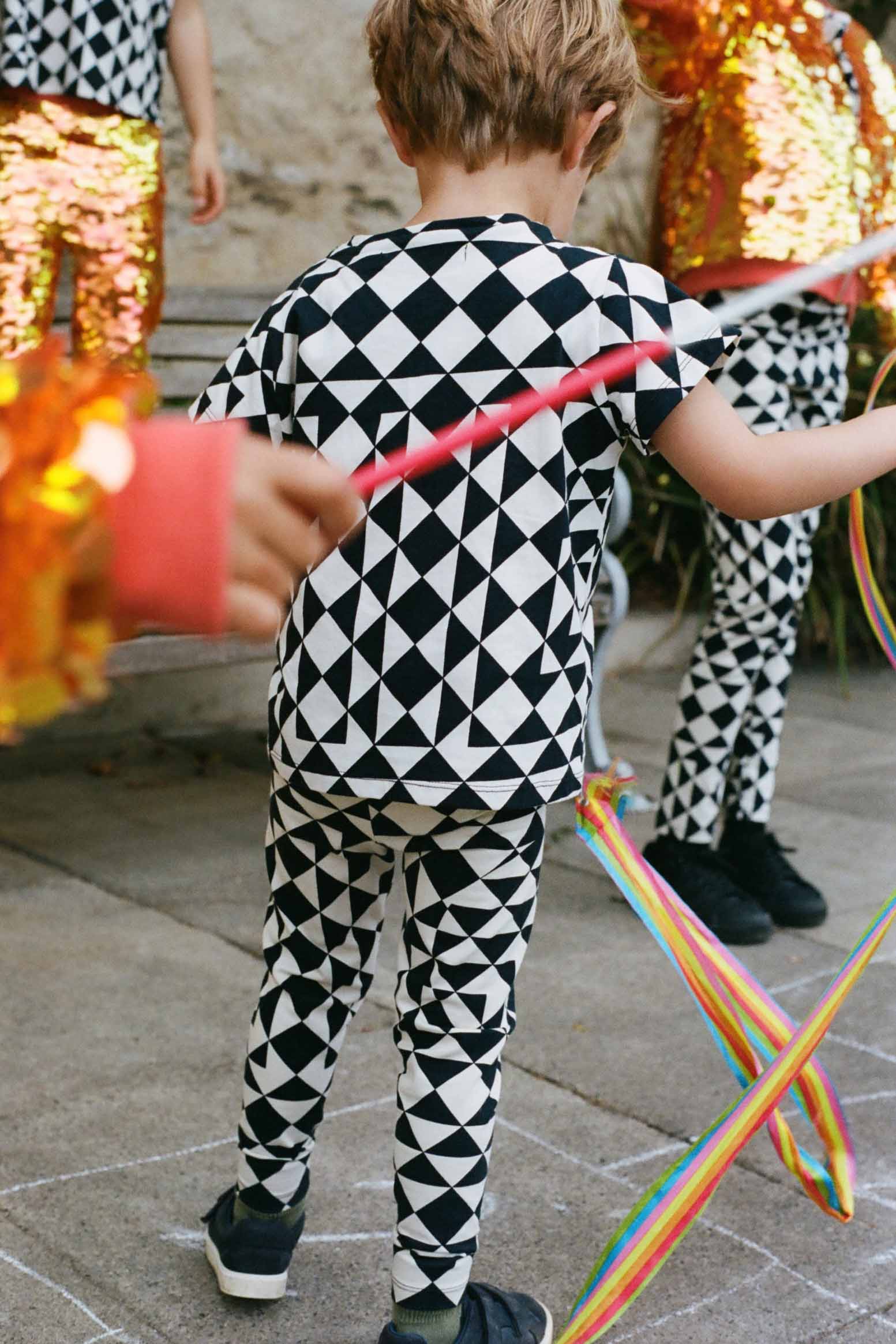 A cropped image of a small child who is standing with their back to the camera playing with swirling colourful ribbons wearing black and white geometric leggings in an outdoor party setting. Cropped from the image is older children wearing kids sequin festival and party leggings and sequin shimmering  bomber jackets. The small boy is wearing black and white geometric  childrens circus/festival patterned cotton leggings in the Rosa Bloom tri-print design with a matching childrens t-shirt. 