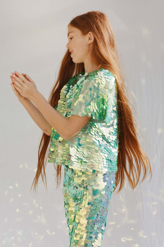 A girl with long hair in a brightly lit outdoor space playing a playground game with their hands. She is wearing Rosa Bloom chameleon mint green, shimmering sequin leggings for children with matching chameleon sequin t-shirt.