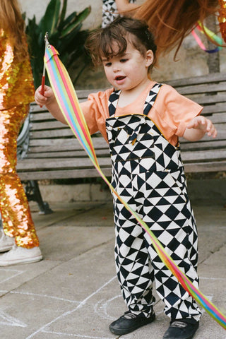 A cropped image of a small child with short dark hair who is wearing black and white tri-print dungarees in the Rosa Bloom tri-print design with a peach coloured childrens t-shirt. Cropped from the photo are two older children wearing matching childresn sequin leggings, playing with brightly coloured ribbons.