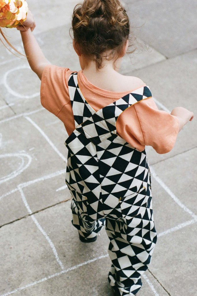A cropped image of a small child with short dark hair who is holding a hand. She is wearing black and white tri-print dungarees in the Rosa Bloom tri-print design with a peach coloured childrens t-shirt.