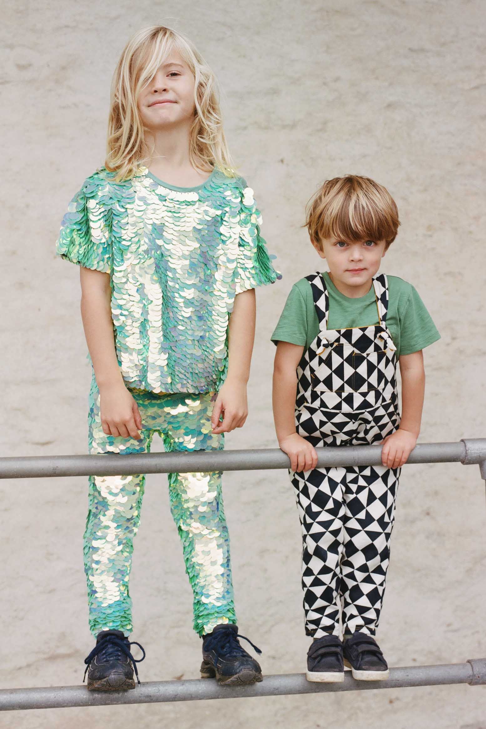 Two children who look like they are having fun in an outdoor park setting, standing on some railings. One child is wearing a mix of chameleon mint green large round sequin festival kids t-shirts and matching leggings. The smaller child is wearing a black and white geometric pair of childrens festival dungarees in a black and white geometric tri-print design by Rosa Bloom. 