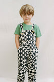 A child wearing a bold, graphic, black and white geometric pair of childrens festival Rosa Bloom dungarees in the Tri-print design, with a contrasting short sleeve green t-shirt by Rosa Bloom. 