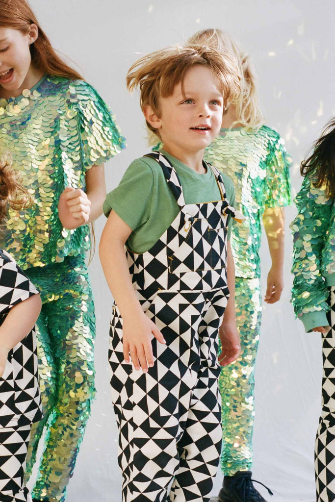 A child jumping up and down in a bold, graphic, black and white geometric pair of childrens festival Rosa Bloom dungarees in the Tri-print design, with a contrasting short sleeve green t-shirt by Rosa Bloom. He is surrounded by other children who are playing in the background wearing Rosa Bloom childrens wear sequin outfits. They are wearing the chameleon dreamy iridescent sequin leggings and matching sequin t-shirts for children. 
