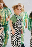 A child jumping up and down in a bold, graphic, black and white geometric pair of childrens festival Rosa Bloom dungarees in the Tri-print design, with a contrasting short sleeve green t-shirt by Rosa Bloom. He is surrounded by other children who are playing in the background wearing Rosa Bloom childrens wear sequin outfits. They are wearing the chameleon dreamy iridescent sequin leggings and matching sequin t-shirts for children. 