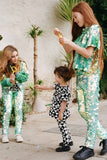 Three children in a brightly lit outdoor walled garden space with tropical plants. Two older children are playing with bubbles with a smaller child. They are all wearing Rosa Bloom chameleon mint green, shimmering sequin clothing. They are wearing the chameleon sequin leggings, sequin childrens t-shirts and matching festival sequin bomber jacket. The little one is dressed in Childrenswear Rosa Bloom tri-print in black and white.