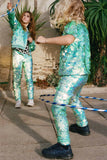 Two children with long hair in a brightly lit outdoor walled garden. The two children are having fun playing with hula hoops like in a circus show. They are both wearing Rosa Bloom chameleon mint green, shimmering sequin childrens t-shirt and matching festival sequin childrens Luna design leggings. 