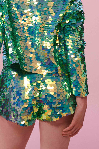 A rear view of a woman wearing iridescent mint green Juno high waisted, tailored sequin shorts covered in large round holographic Rosa Bloom sequins. The sequins glisten, creating a mix of shimmering colours of greens that make this sequin shimmer. The model is also wearing a matching stretchy sequin jacket, in matching colours. 