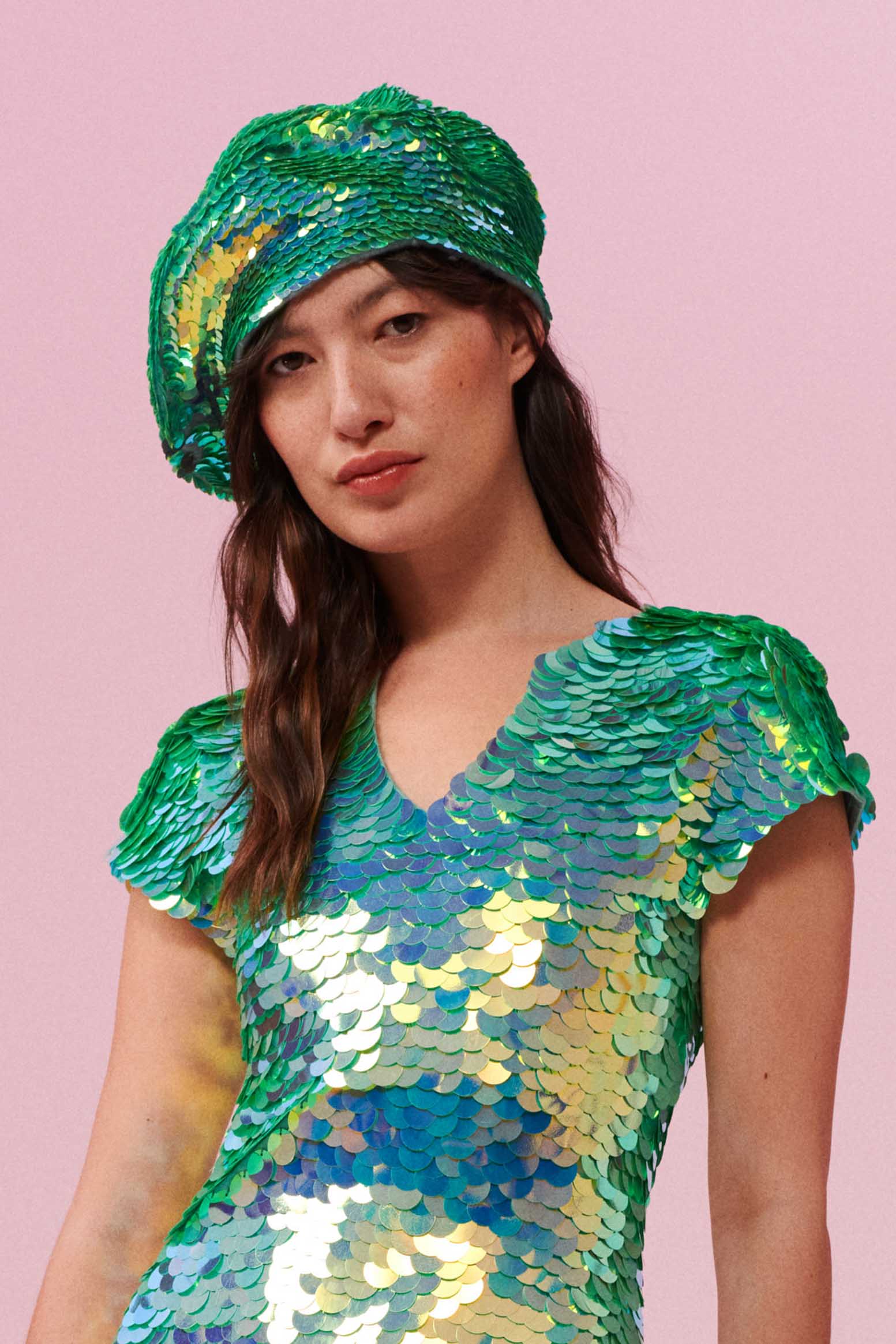 A woman with brown hair swept to the side, wearing a sequin beret made from round holographic sequins with hues of mint and sage green. The festival, woollen and sequin hat matches the sequin top that the model is wearing. The chameleon sequins glisten, creating a mix of shimmering colours.