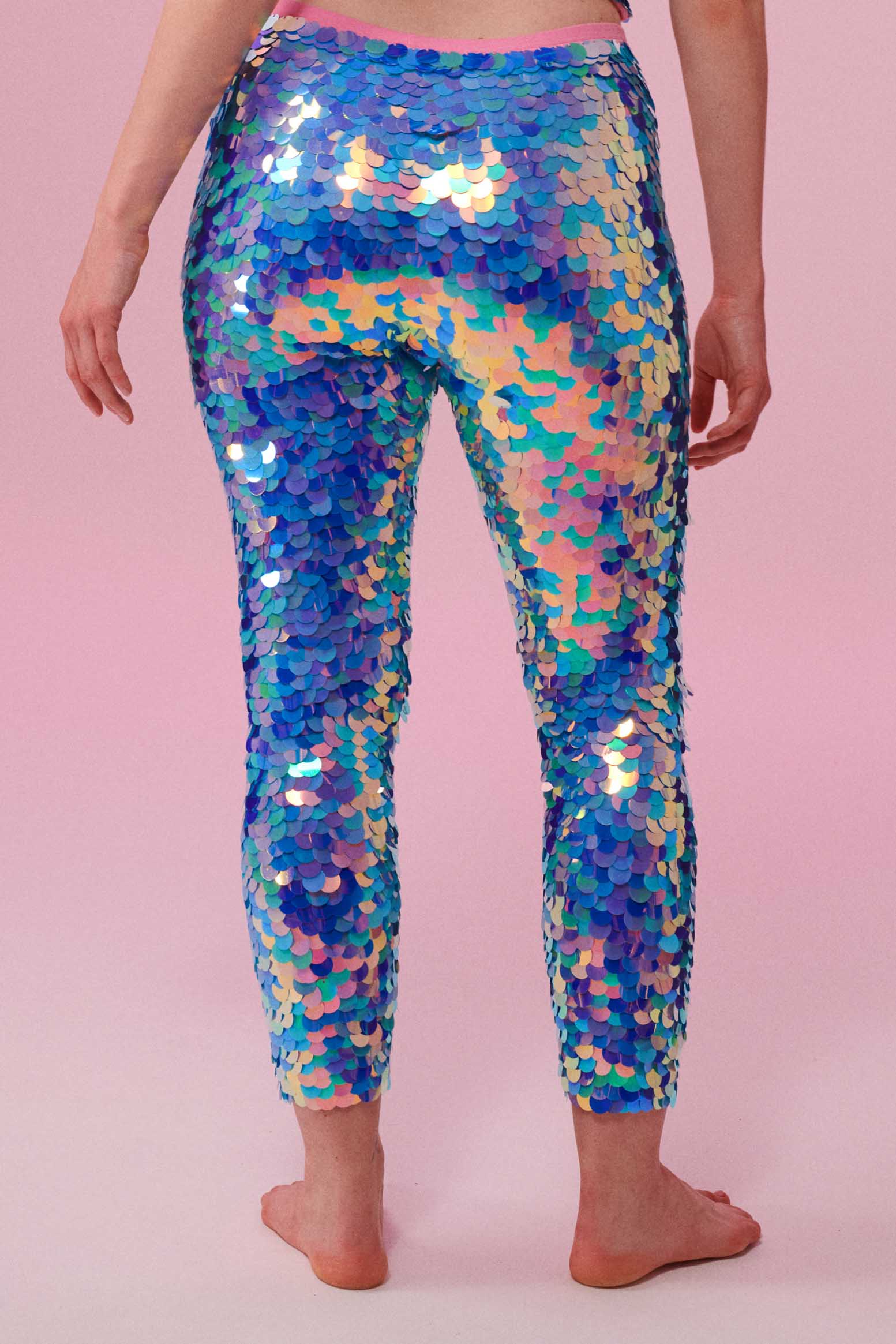 A woman standing with her back to you, wearing iridescent pink, purple and blue, stretchy sequin Indus leggings covered in large round holographic Rosa Bloom sequins. The sequins glisten, creating a mix of shimmering colours  to create this Amethyst leggings design that looks like they are glowing. 