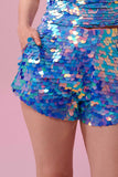 A crop of a woman’s torso wearing iridescent purple, pink, blue and white Juno sequin shorts covered in large round holographic Rosa Bloom sequins. The sequins glisten, creating a mix of shimmering colours make this sequin shimmer. The model is also wearing a matching stretchy sequin top, in matching colours. This Amethyst sequin outfit looks like it is glowing in the light. 