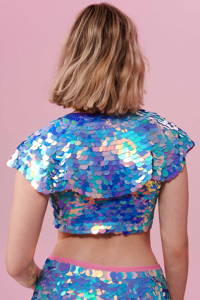 A rear view of a woman with short blonde hair, wearing a festival sequin cape made with small and large round holographic Rosa Bloom sequins. The Amethyst sequins, sitting neatly over her shoulders in this Inti design glistens in the light, creating a mix of shimmering colours of purple, pink, blue and white. 