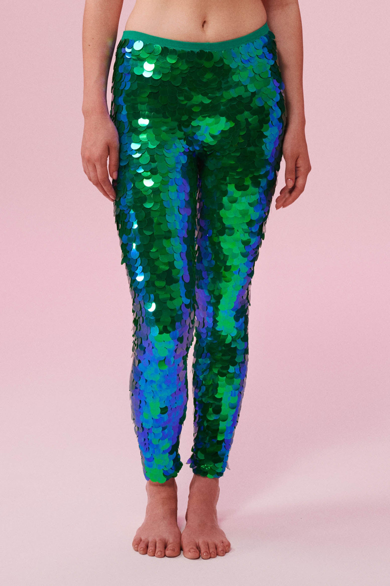 A crop of a woman’s legs wearing iridescent blue and green Indus leggings covered in large round holographic Rosa Bloom sequins. The sequins glisten, creating a mix of shimmering colours make this sequin glow. The model is also wearing a matching stretchy sequin vest top, in matching colours. This Emerald sequin outfit looks like it is glowing. 