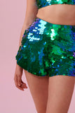 A crop of a womans torso wearing iridescent green and blue Juno high waisted, tailored sequin shorts covered in large round holographic Rosa Bloom sequins. The sequins glisten, creating a mix of shimmering colours of greens and blues. The model is also wearing a matching stretchy sequin cropped vest, in matching colours. 