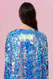A woman with long brown hair swept over her shoulder standing with her back to the camera in a brightly lit indoor room with pink walls wearing a Rosa Bloom blue and pink sequin jacket