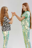 Two children in a brightly lit outdoor space playing a playground game with their hands. They are wearing Rosa Bloom chameleon mint green, shimmering sequin leggings for children. One girl is wearing the matching chameleon sequin t-shirt, and the other is wearing a geometric, bold and graphic black and white Rosa Bloom tri-print t-shirt. 