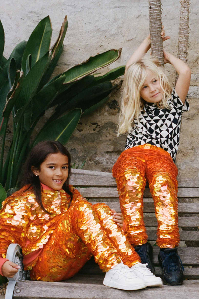 Two children in a brightly lit outdoor walled garden space with tropical plants, having fun sitting on a bench. They are both wearing Rosa Bloom fox - dreamy orange and yellow, shimmering sequin clothing. They are wearing the fox sequin leggings for children, with matching festival sequin bomber jacket. The other child is wearing a childrenswear Rosa Bloom bold graphic tri-print t-shirt in black and white.