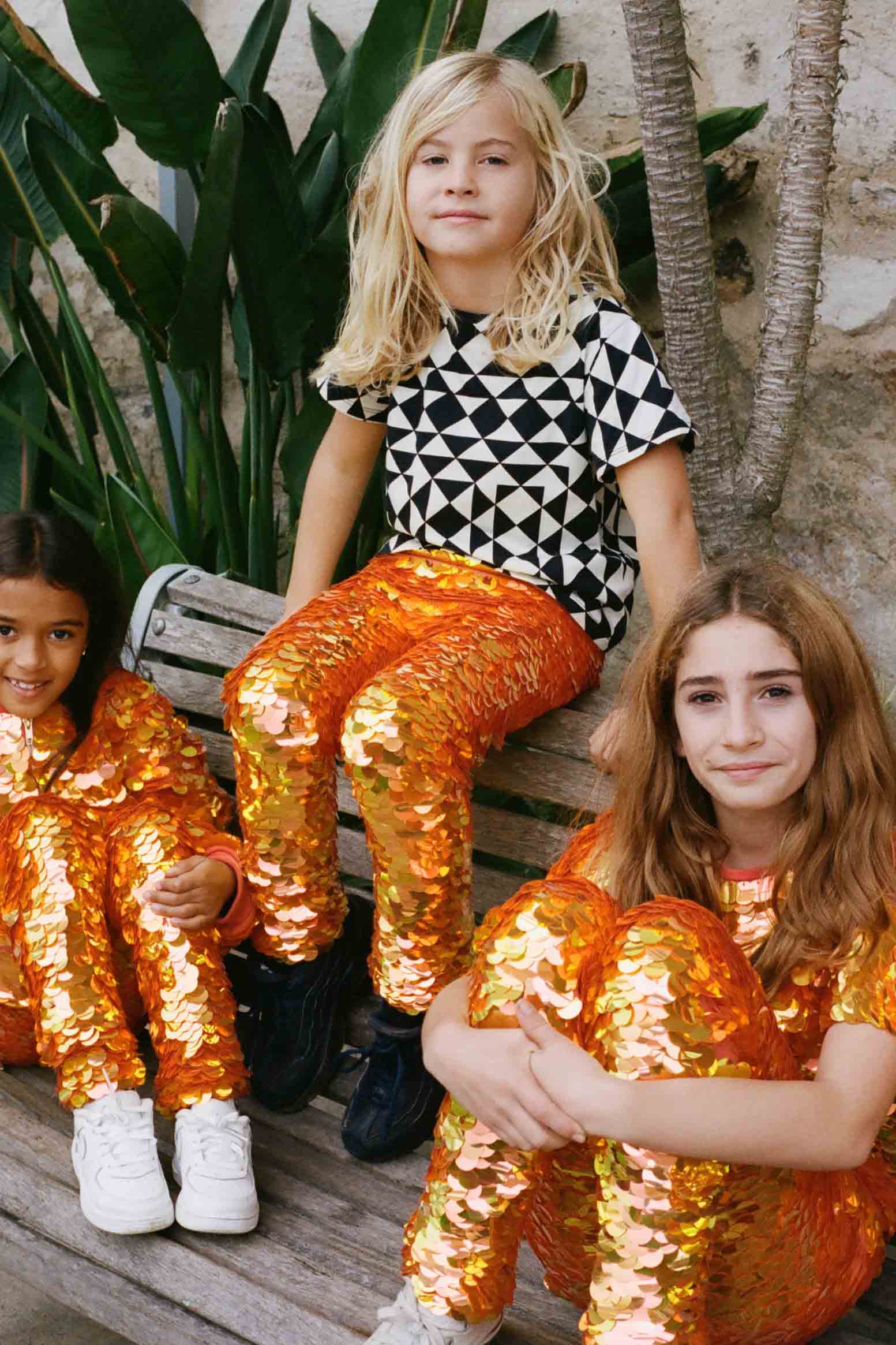 Three children in a brightly lit outdoor walled garden space with tropical plants, sitting on a bench, posing for the camera. They are all wearing Rosa Bloom fox - dreamy orange and yellow, shimmering sequin clothing. They are wearing the fox sequin leggings for children, matching sequin childrens t-shirts and matching festival sequin bomber jacket. The child in the middle is wearing a Childrenswear Rosa Bloom bold graphic tri-print in black and white.