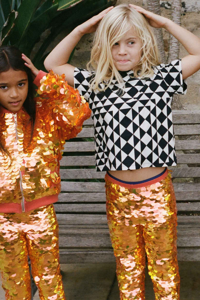 Two children in a brightly lit outdoor walled garden space with tropical plants, having fun playing playground games. They are both wearing Rosa Bloom fox - dreamy orange and yellow, shimmering sequin clothing. They are wearing the fox sequin leggings for children, with matching festival sequin bomber jacket. The other child is wearing a childrenswear Rosa Bloom bold graphic tri-print t-shirt in black and white.