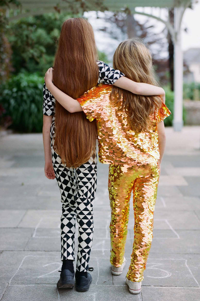 Two children with long hair with their backs to us in an outdoor park setting. The two children have their arms around each other. One is wearing a fox orange and yellow shimmering sequin festival kids t-shirt and leggings. The other is wearing a black and white geometric and triangle pair of Childrens leggings with a matching short sleeve black and white geometric tri-print t-shirt. 