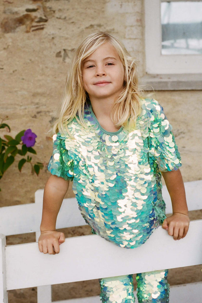 A child in a brightly lit outdoor walled garden space with tropical plants, standing on a white fence looking at the camera. He is wearing Rosa Bloom chameleon mint green, shimmering sequin childrens wear. He wearing the chameleon sequin leggings and matching sequin childrens t-shirt. 