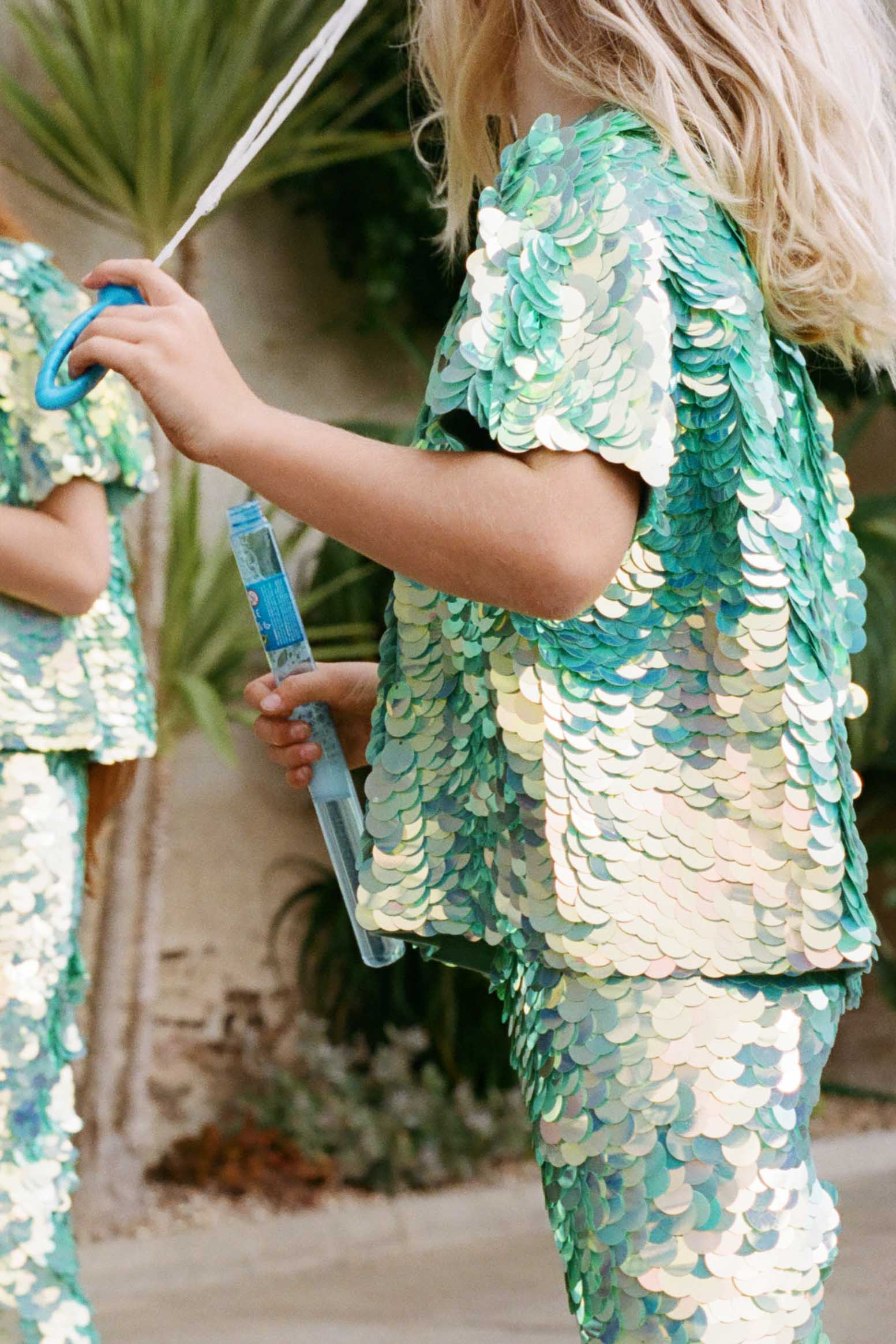 A cropped image of a child in a brightly lit outdoor walled garden space with tropical plants, splaying with bubbles. He is wearing Rosa Bloom chameleon mint green, shimmering sequin childrenswear. He wearing the chameleon Luna sequin leggings and matching sequin Sonny children’s t-shirt. 