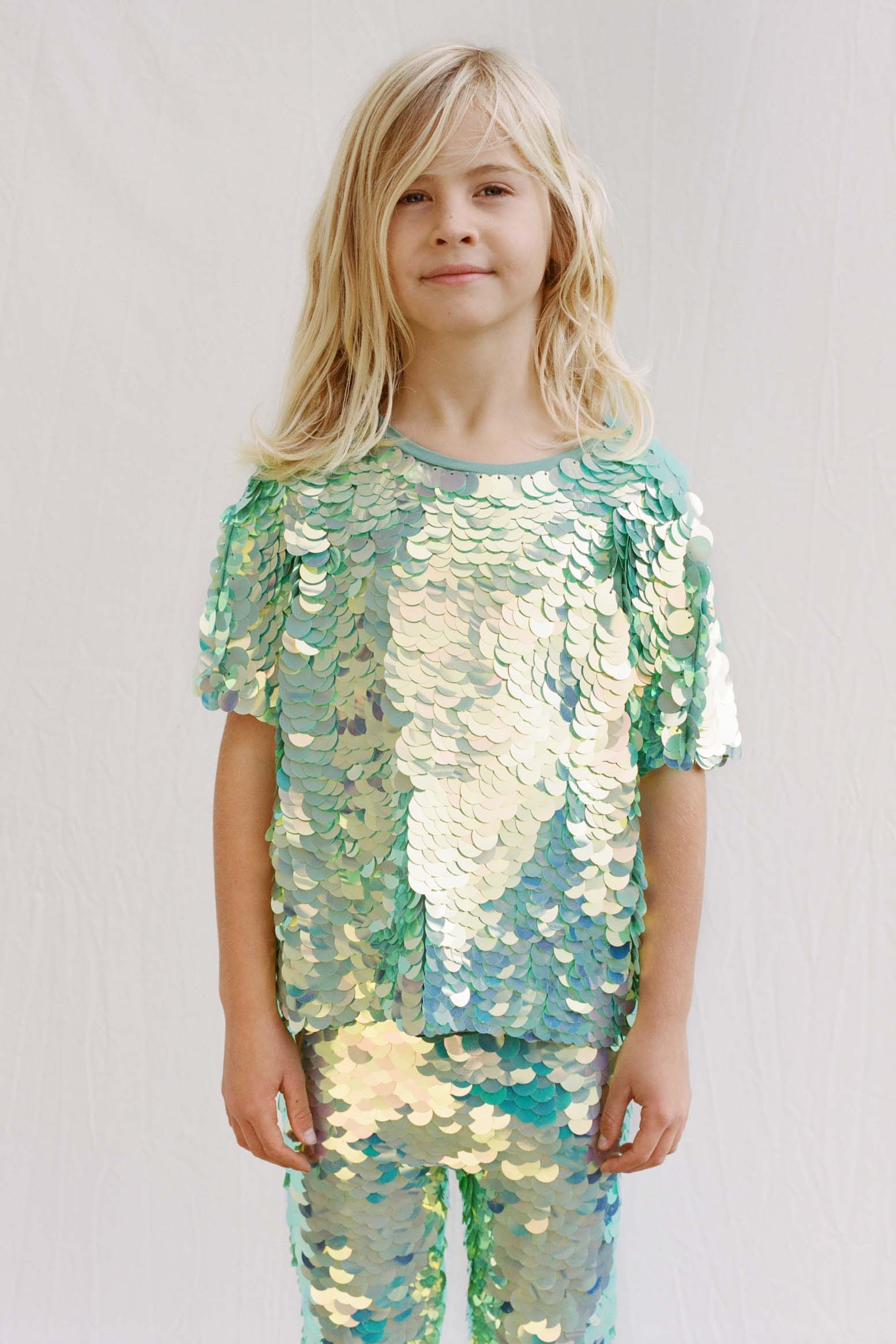 An image of a child in a brightly lit outdoor space, facing the camera. He is wearing Rosa Bloom chameleon mint green, shimmering sequin childrenswear. He wearing the chameleon Luna sequin leggings and matching sequin Sonny children’s t-shirt. 