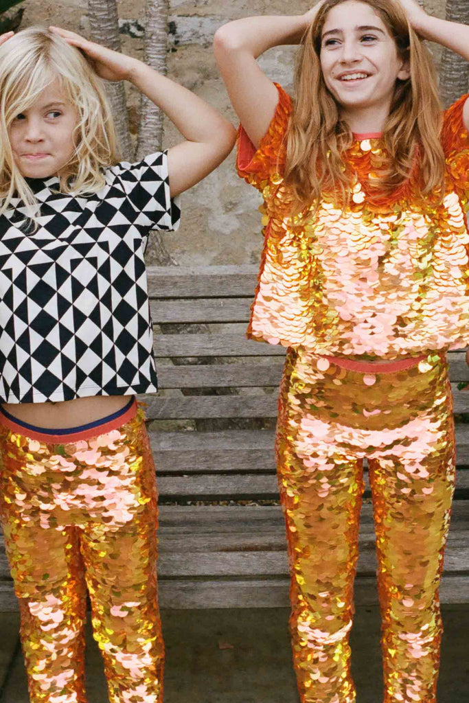 Two children who look like they are having fun in an outdoor park setting, standing in front of a park bench. They are wearing a mix of orange, gold and yellow large round sequin festival kids childrenswear in leggings and t-shirt, and a geometric graphic black and white Tri-print t-shirt by Rosa Bloom. 