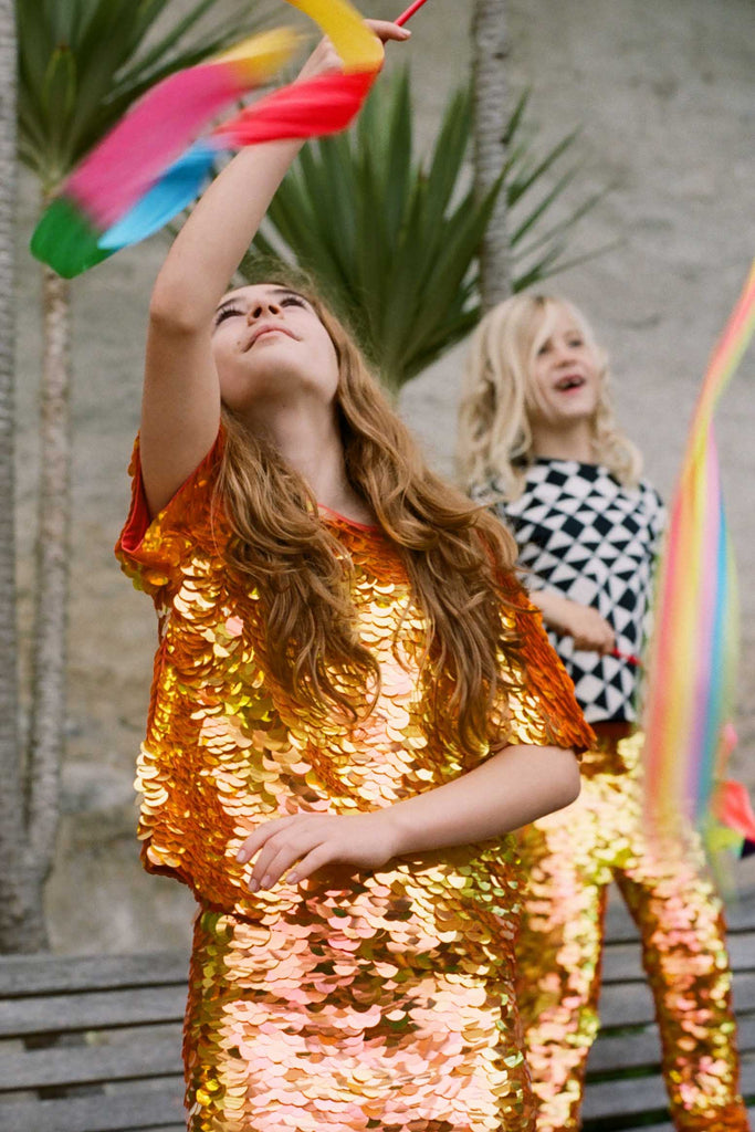  An image of a child in a brightly lit outdoor festival circus space, playing with bright coloured flowing ribbon. She is wearing Rosa Blooom Sonny T-shirt in fox colourway - shimmering sequin childrenswear. 