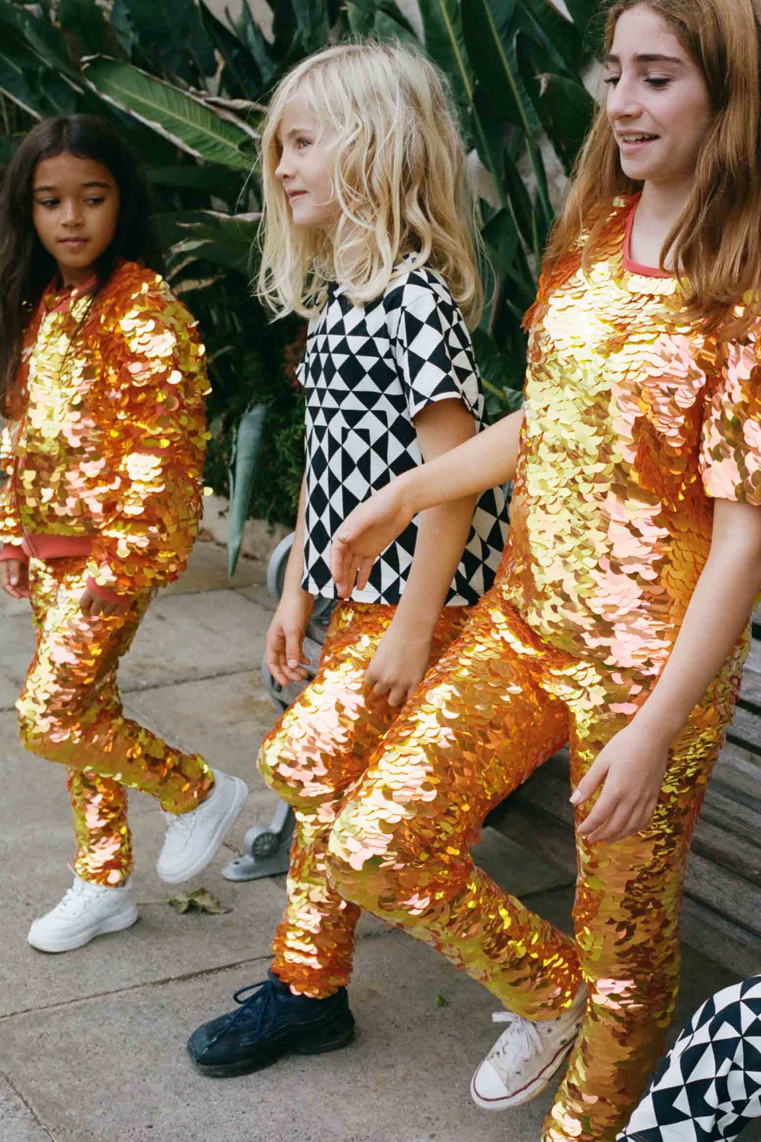 Three children in a brightly lit outdoor walled garden space with tropical plants. They are all wearing Rosa Bloom fox m orange, gold and yellow, shimmering sequin clothing. They are wearing the fox sequin leggings, sequin childrens t-shirts and matching festival sequin bomber jacket. The middle child is dressed in Childrenswear Rosa Bloom tri-print in black and white.