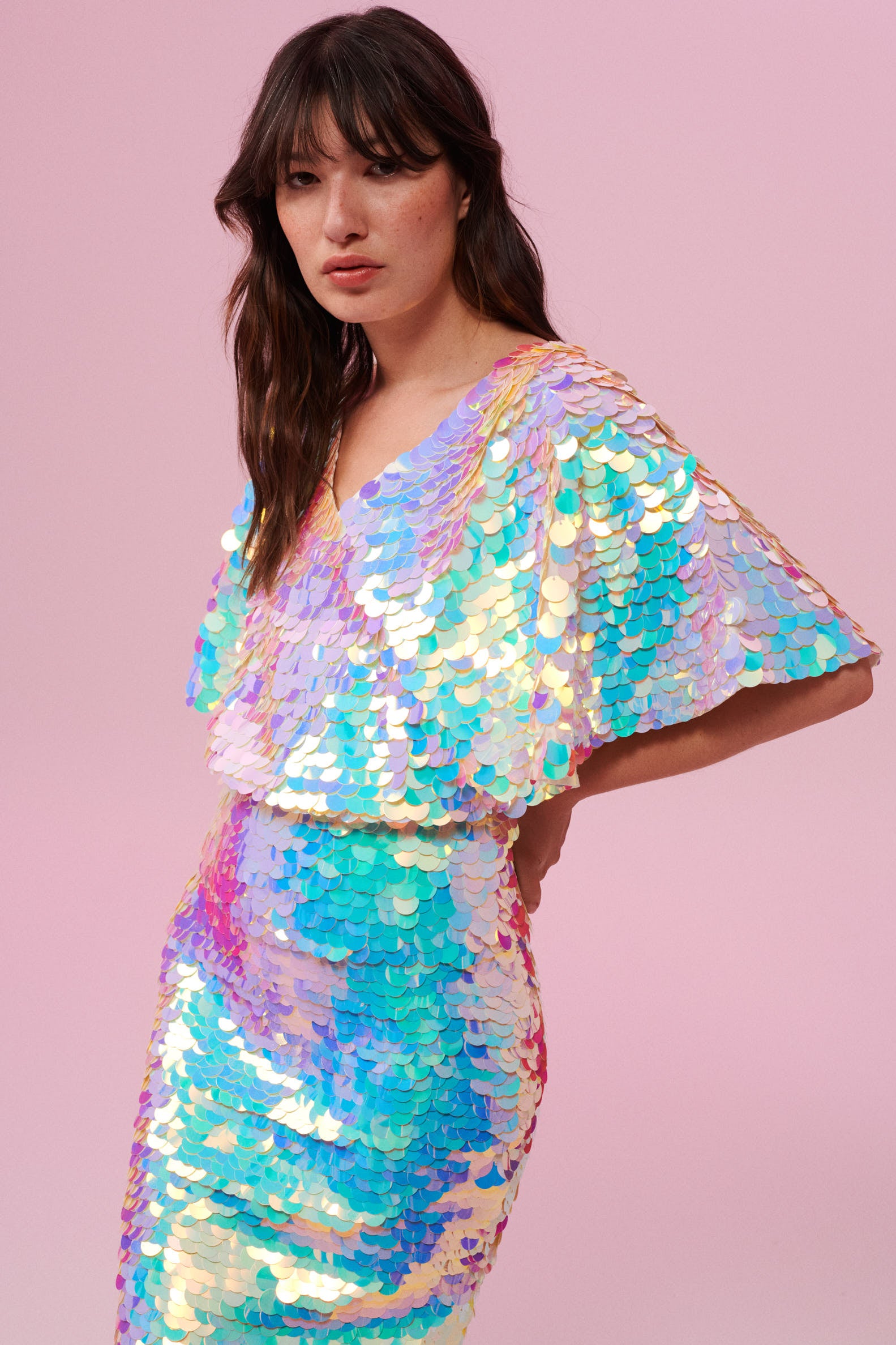 A woman facing towards you at an angle wearing an opal sequin top with large cape sleeves covered in large round holographic purple,pink, blue and white coloured Rosa Bloom sequins.